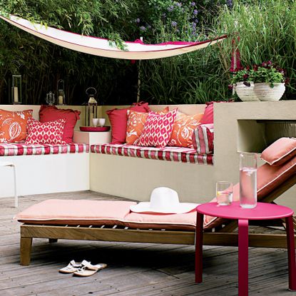 A garden with a wooden adjustable recliner sun lounger with a pik cushion pad and built-in bench seating adorned with patterned pink cushions