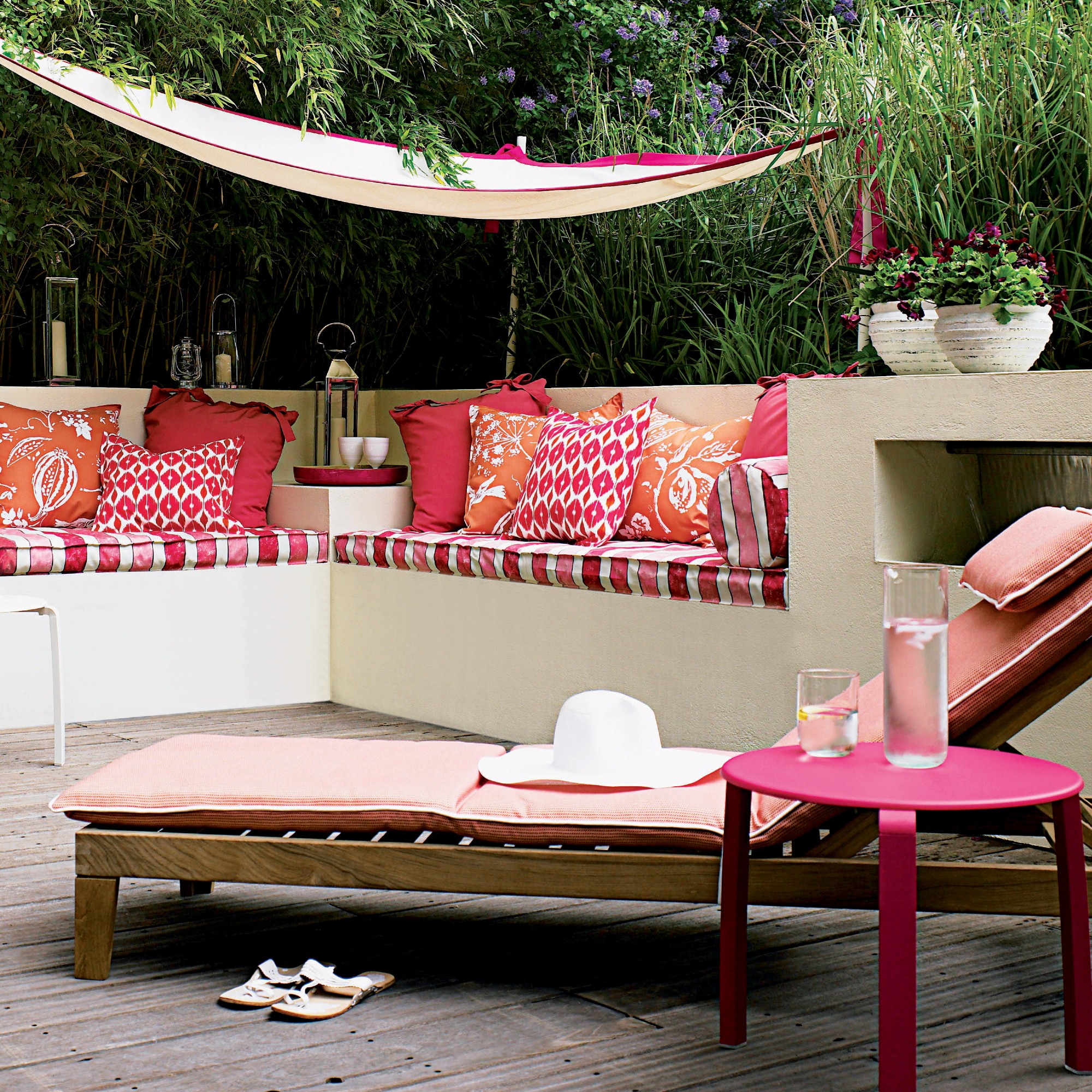 A garden with a wooden adjustable recliner sun lounger with a pik cushion pad and built-in bench seating adorned with patterned pink cushions