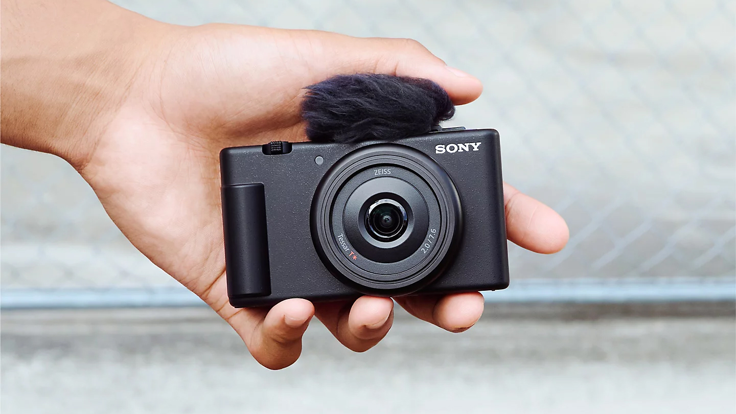 A hand holds a compact SonyZv-1F camera with a furry windscreen attached to the microphone, set against a blurred background