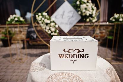 A wooden box with the word "wedding" on it ready to collect cards of gifts from guests, with a floral backdrop in the background.