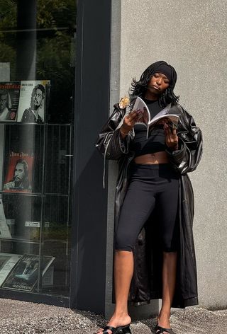 A woman wearing a long black leather trench coat over a black cropped t-shirt, black capri pants, and black mules.