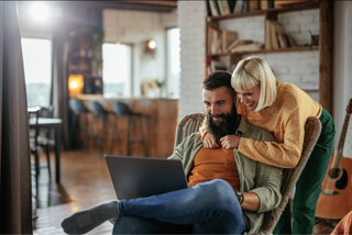A cheerful couple in a cozy room, with a man on a laptop and a woman affectionately leaning over him.