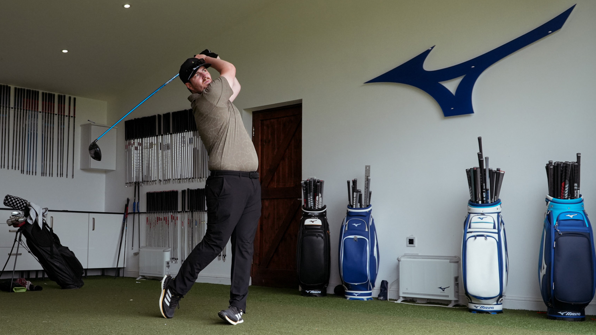 A golfer holds their finish in a hitting studio with a Mizuno logo on the wall