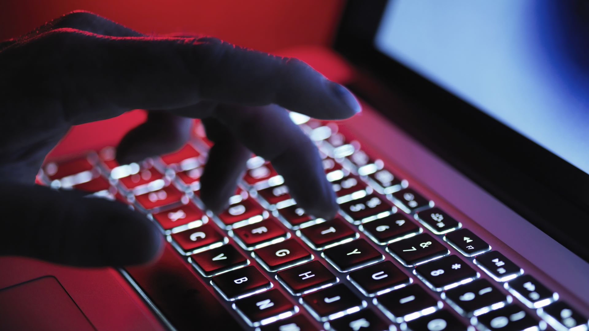 A hand typing at a computer in a dark room, lit up by the laptop's keyboard LEDs and red LED light