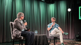 Chris Dring interviews Xbox Chief Strategy Officer Matthew Ball on stage during a live event, with both seated in armchairs holding microphones under green lighting.