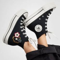 Woman's feet in air wearing black Converse Chuck Taylors with embroidered flowers.