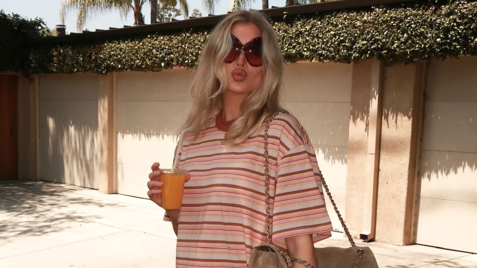 Elsa Hosk holds an orange juice wearing a striped t-shirt with white jeans and brown flip-flops.