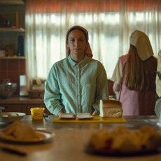 a woman in traditionalist clothing sits at a table slicing bread in a still from netflix's unchosen