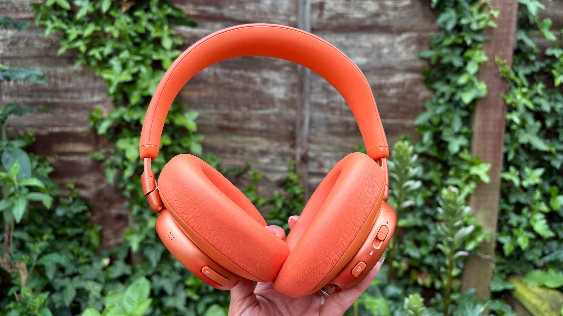JBL Live 780NC wireless headphones held in hand in front of garden fence