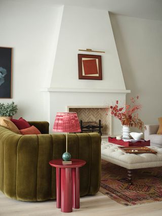 A living room with a green velvet sofa, a fireplace, and a pink pedestal side table with a pink table lamp on it