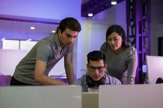 Three software engineers watching a laptop screen, with one sat down at a desk on the laptop and the other two leaning on the desk to either side of him, in an office dimly-lit with purple light