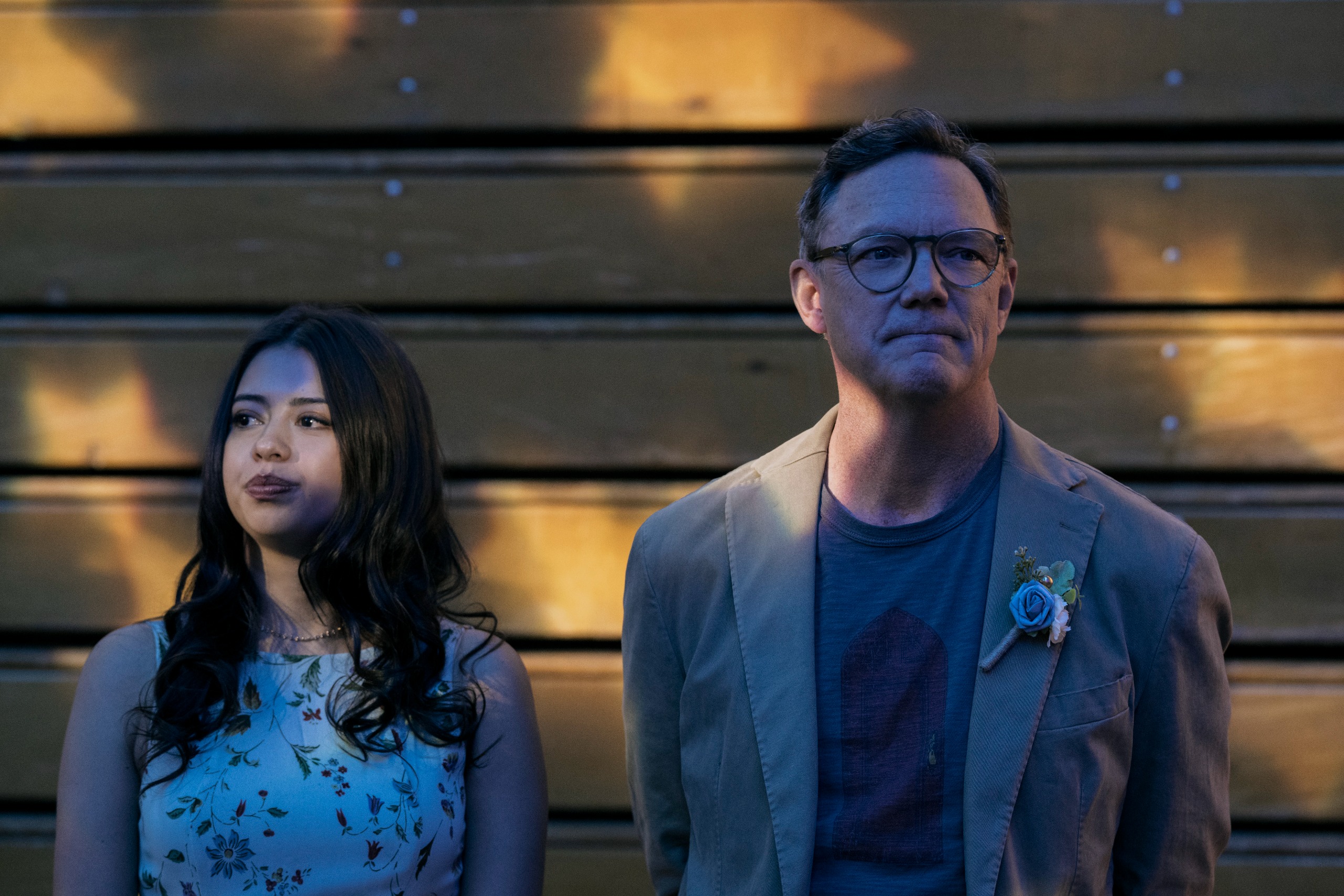 A woman and man in formal wear (Amber Midthunder as Rita Desjardin and Matthew Lillard as Henry Grayle) stand in front of a wooden wall during a high-school dance, in Prime Video's 'Carrie.'