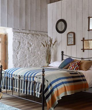 A farmhouse bedroom with an iron bedframe with a striped throw and throw pillows on the bed, distressed white wall panels and a gallery wall of mirrors above the bed