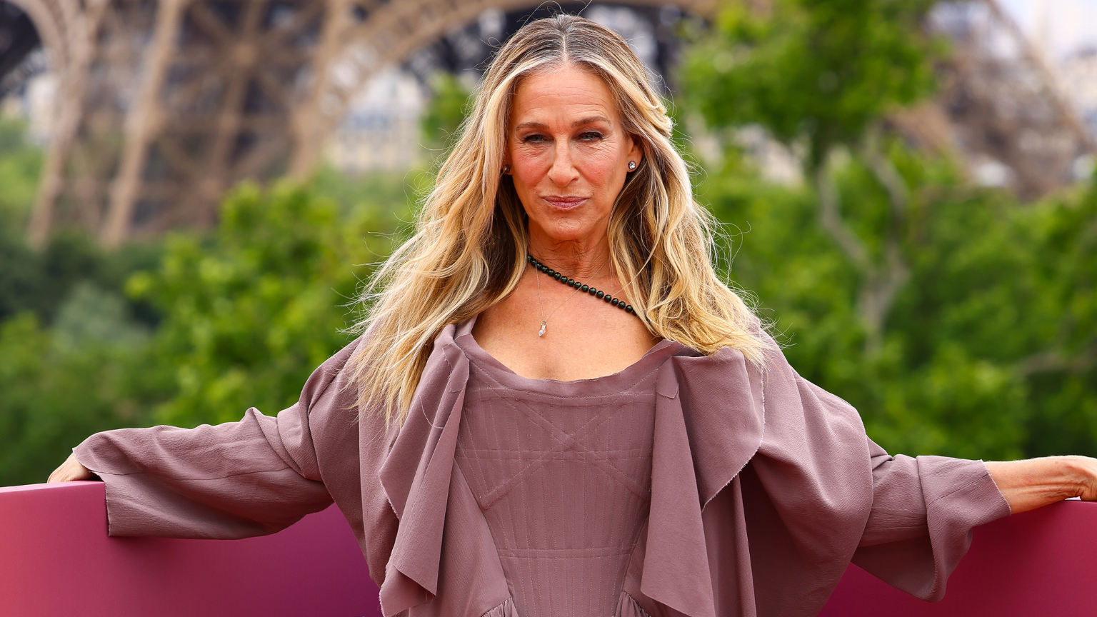 a woman with long blonde hair wearing a mauve dress in front of trees and a pink wall