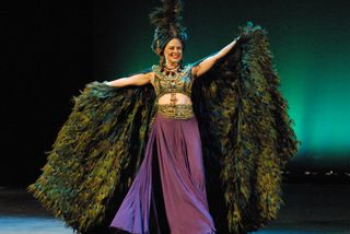 Smiling female holding out a cape of peacock feathers, wearing a long purple skirt and green and gold jewelled top, green velvet headdress with peacock feathers protruding out of the top, area behind her is la green wall lit up with spotlighting