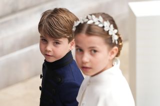 Princess Charlotte wears a white dress and a floral headband while Prince Louis wears a navy jacket while he stands behind her