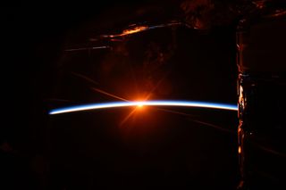 a bright, blue-white arch stretches across the center from the left, a bright yellow point of light shines at its center. A partially illuminated section of spacecraft hangs from above, glowing faint orange.