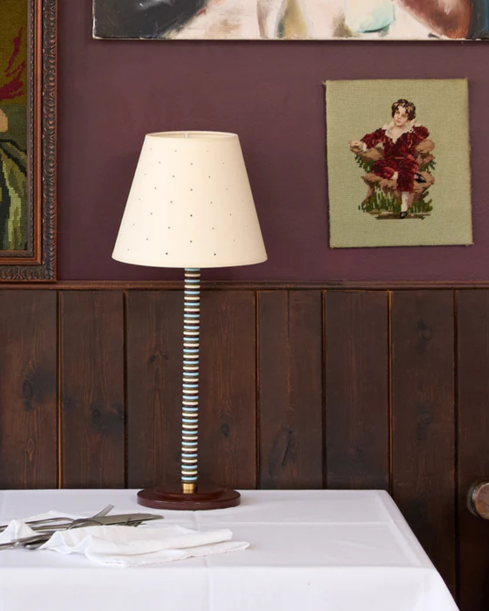 Image of a wall that is plum purple with dark wood paneling on the lower half. There are three framed pictures in view and a striped table lamp with a beige pinhole lampshade.