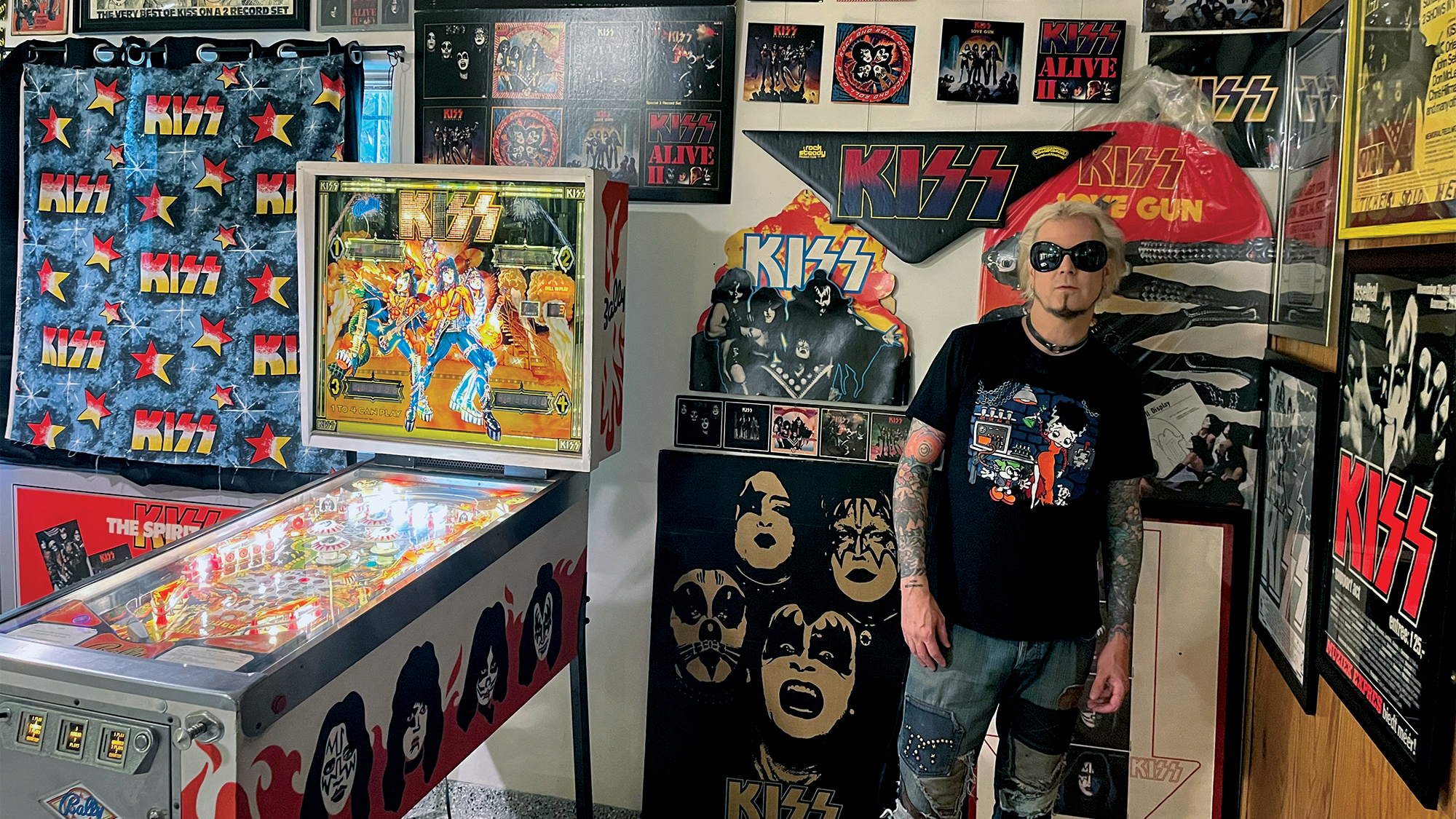 John 5 poses in his Knights in Satan's Service Museum of Kiss Memorabilia. John stands next to a 1979 Bally Kiss pinball machine that sits among rare promotional in-store mobiles and standups. 