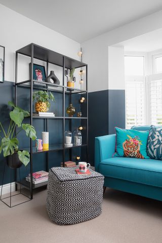Living room with half blue, bright blue velvet sofa, and black shelving unit dressed with plants, vases and other ornaments