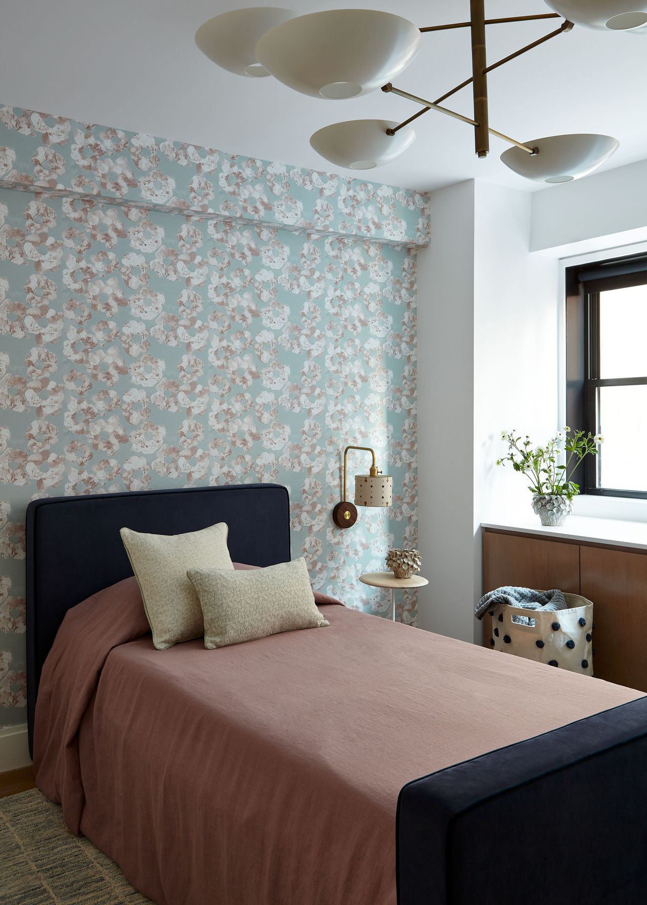 A girl's bedroom with a single bed with a dark blue bedframe and dusty pink bedding, contemporary pendant light, floral wallpaper and a wall sconce next to the small nightstand, a laundry basket next to in-built storage under the windowstill with a plant on top