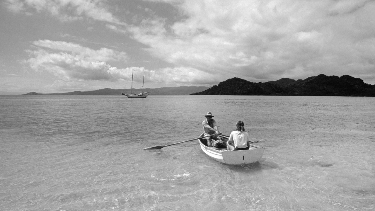 Two people sit in a rowboat near an island in a black and white photo