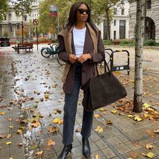 Woman wears grey jeans, a white t-shirt, a brown blazer with a sweater draped over it and black pointy-toe boots. She's carrying a The Row Margaux bag. 