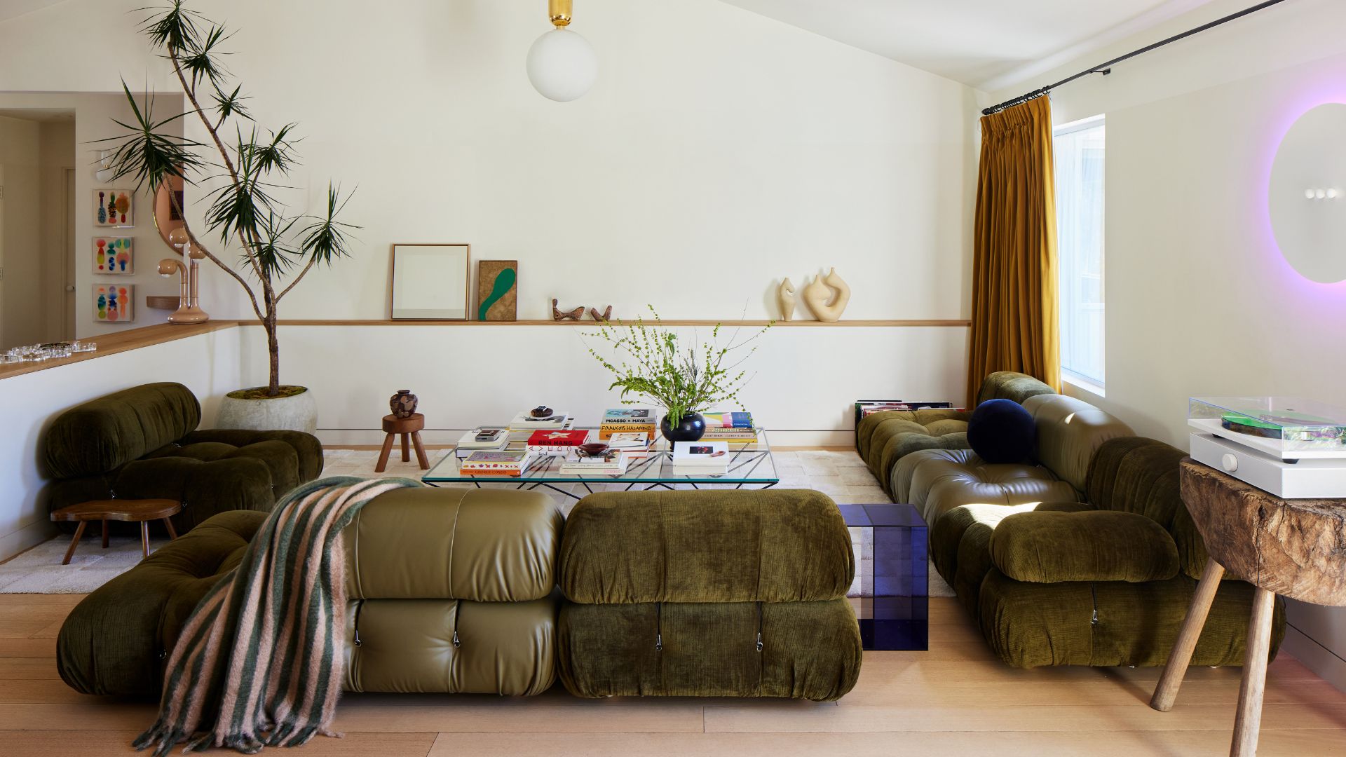 A white living room with a green suede and leather couch , a glass coffee table, a striped throw, wood stools, a record player, and a potted tree