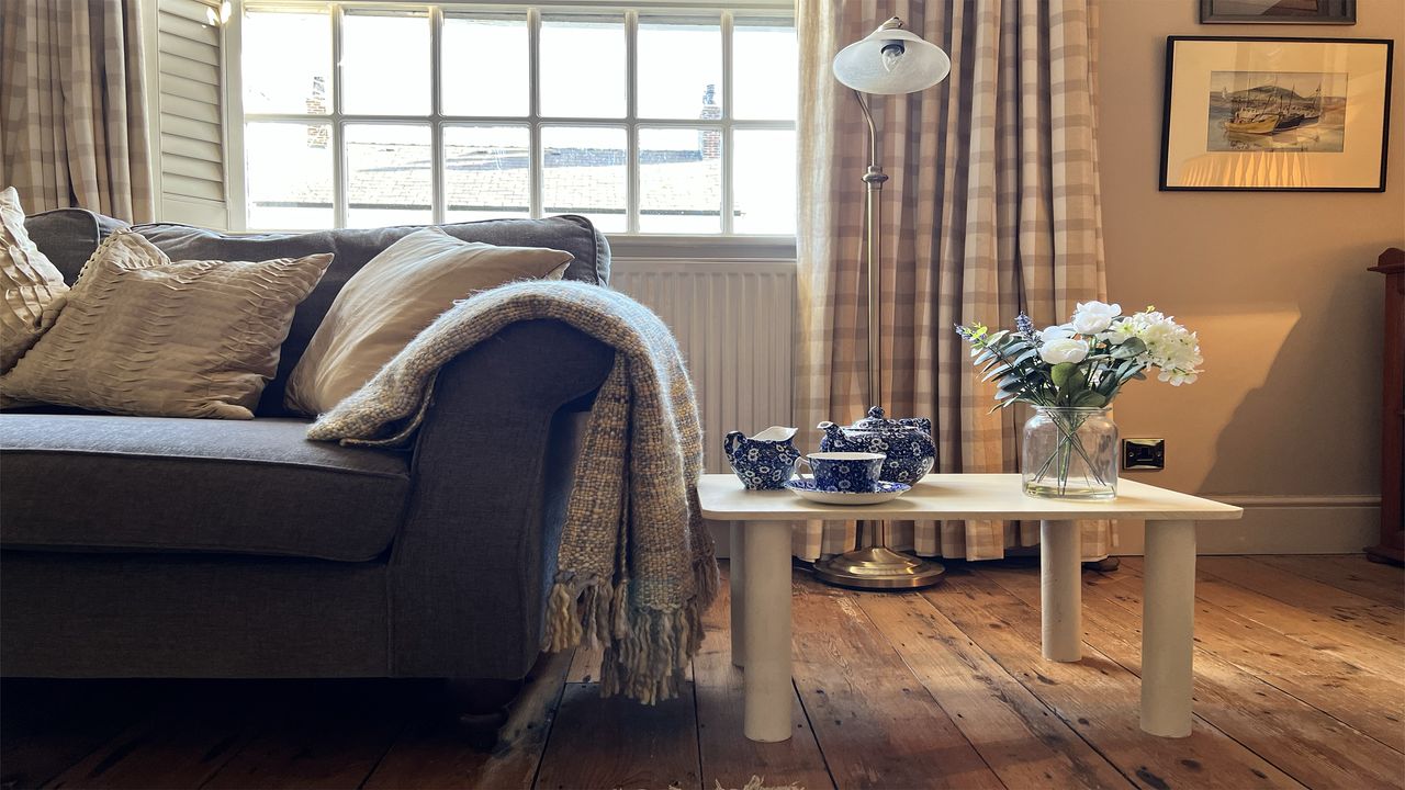 Cream home made table next to sofa with cushions on wooden floor with vase and flower and teapot on top 