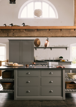 open plan kitchen with grey cabinetry. Large island with integrated hob, above is exposed wooden beams with copper pots hanging off