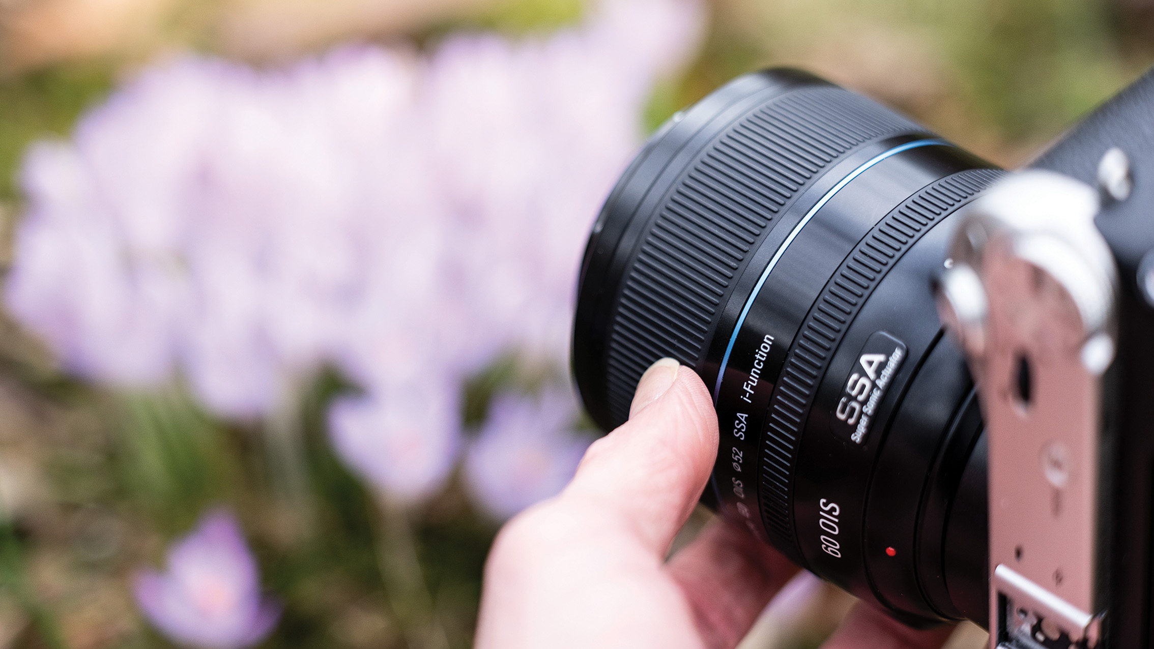 Close-up of a hand holding a camera with a large lens, focused on the lens details. Blurred background of soft purple flowers