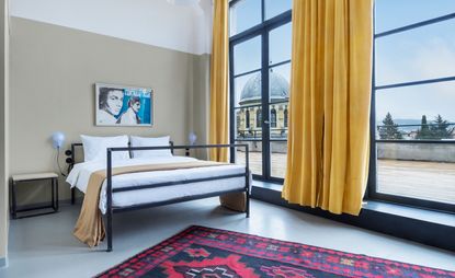 Bedroom showing a bed with white bedding, full-length window with yellow curtains, and a red patterned rug