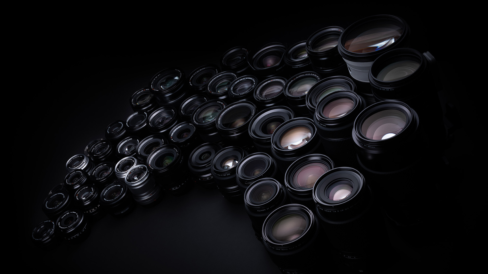 A wide collection of Fujifilm lenses, illuminated with low key lighting, on a black background