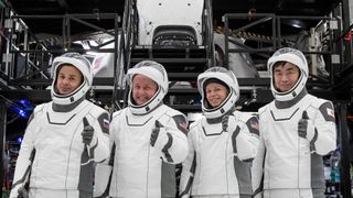 four people in white spacesuits smile and give a thumbs-up in front of a cone-shaped white spacecraft in a hangar surrounded by scaffolding