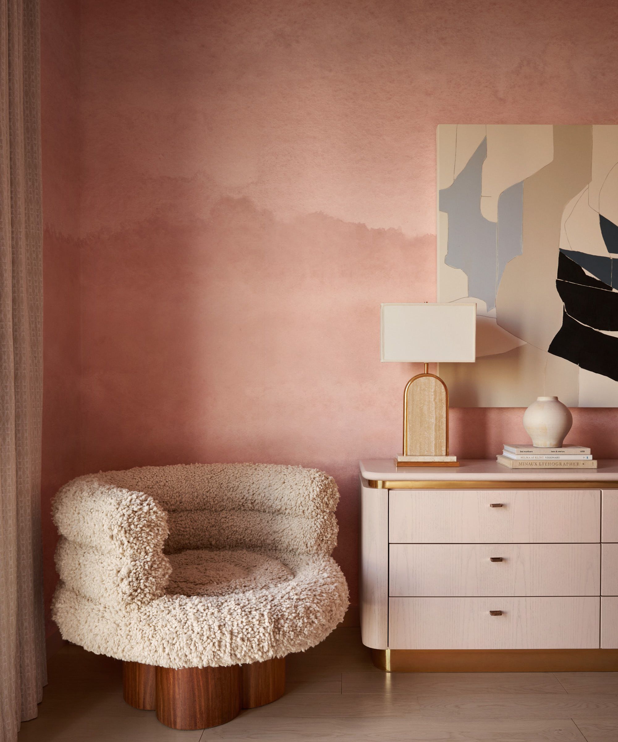 Pink bedroom with light pink console holding lamp and vase, abstract painting and a boucle armchair