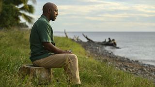 DI Mervin Wilson (Don Gilet) sits on a tree stump on the shoreline, his face intense. Just about visible in his hands are some air-mail letters that he is holding. In front of him is a small, pebbled area of beach and then the sea.