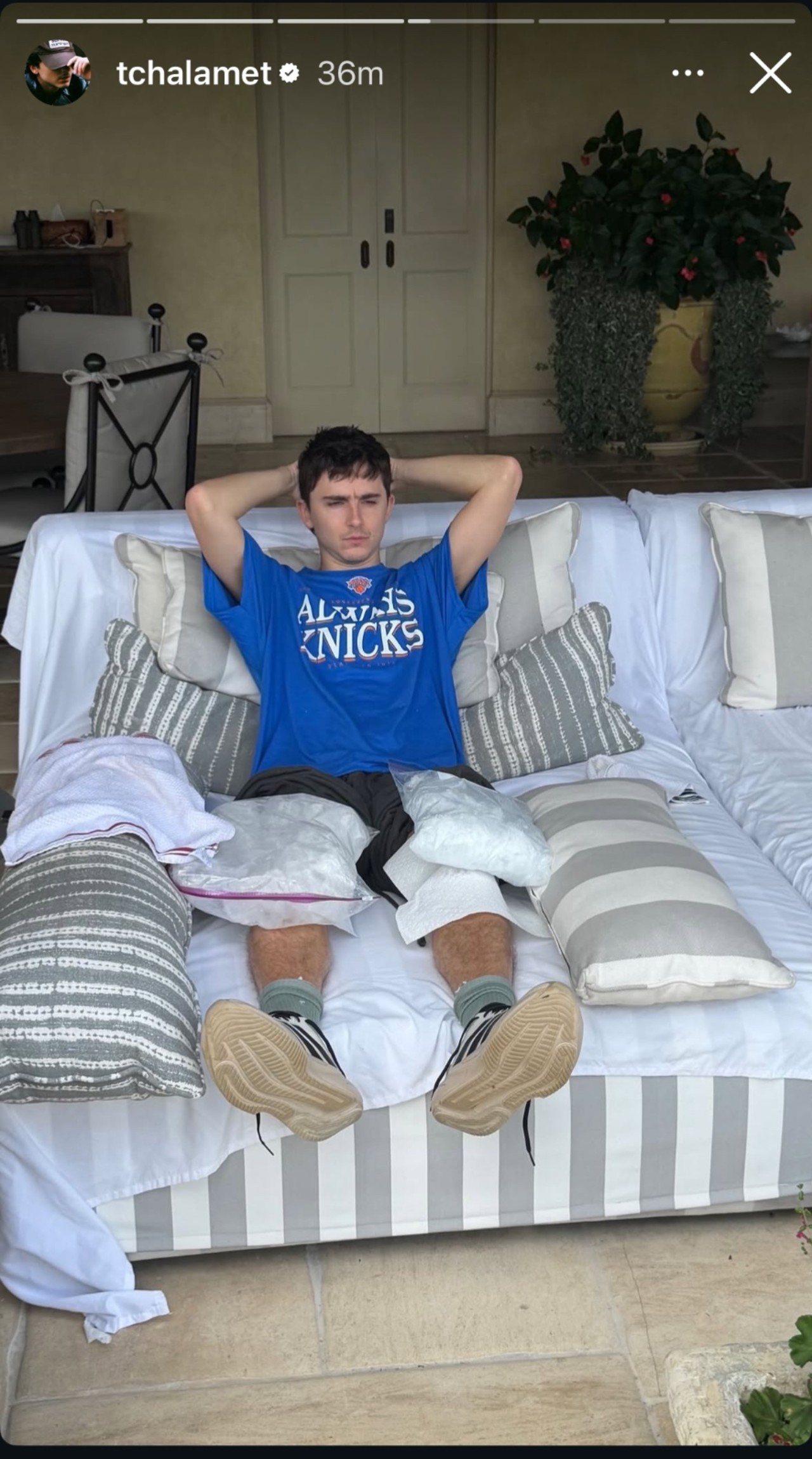Timoth&eacute;e Chalamet sitting on a couch, icing his knees and resting his arms on top of his head. 