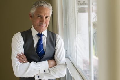 A wealthy-looking businessman in his 50s poses next to a window with his arms crossed.