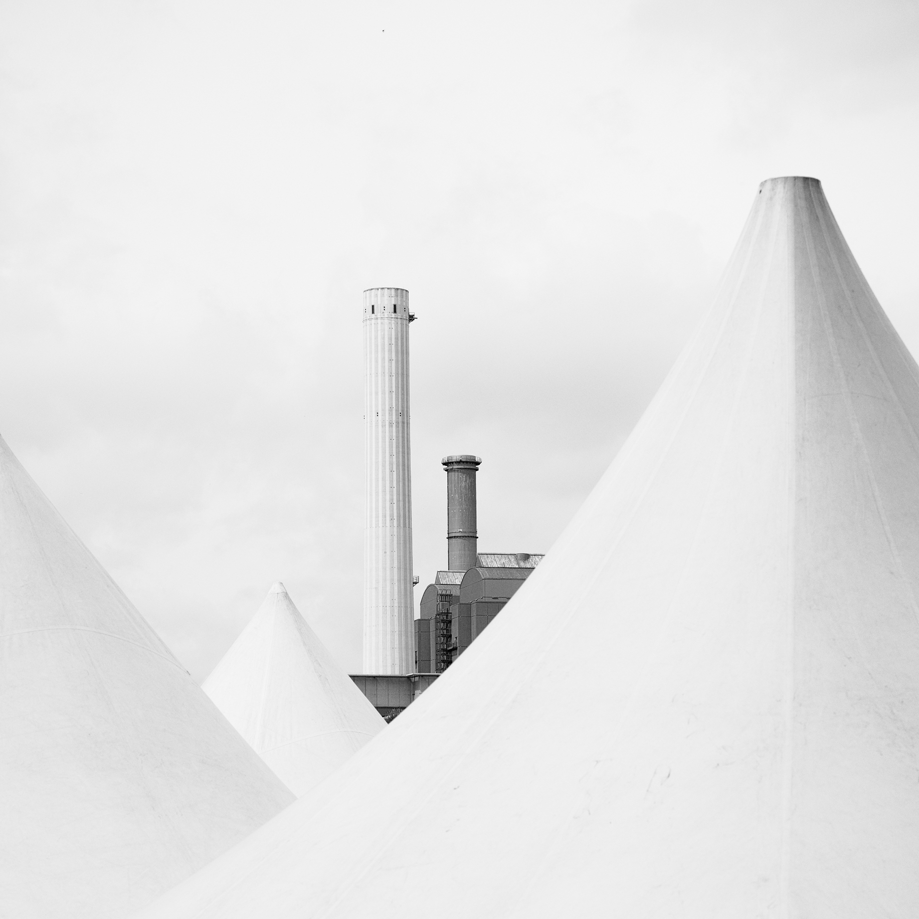A black and white image featuring large, cone-shaped tents in the foreground, set against a backdrop of industrial chimneys under a cloudy sky. The scene feels serene yet industrial