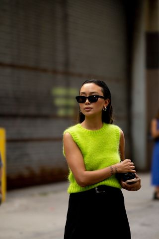 A woman at Paris Fashion Week wearing a gray sweater vest, gray jeans, Dior bag, and black heels.