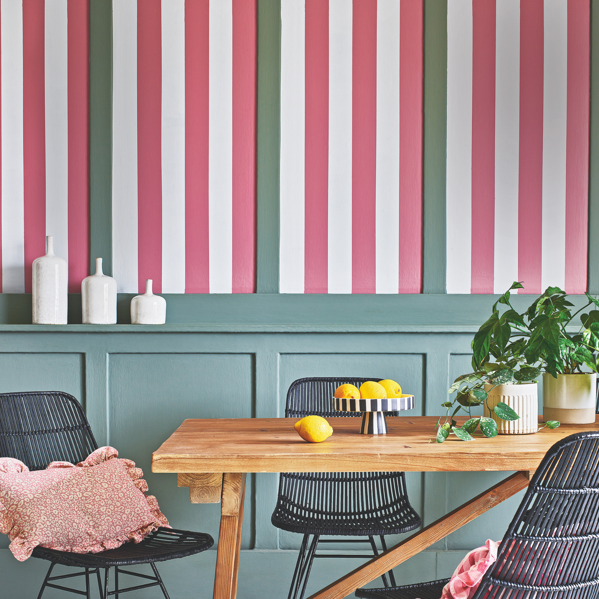 Wood dining table and black woven chairs in front of a pink, white and green painted wall.