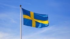 Flag of Sweden flying in the wind with blue skies and light clouds in background.