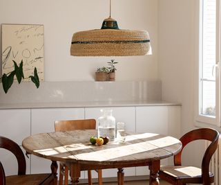 large wicker light shade with green band around middle hanging above round wooden table with chairs, cream handleless kitchen units in background