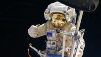 an astronaut in space, his golden visor reflecting a bent image of a space station module out of frame, gives a thumbs up. In the foreground immediately in front of him, a contraption of metal rods, similar to a folded camera tripod, extends the height of the image.