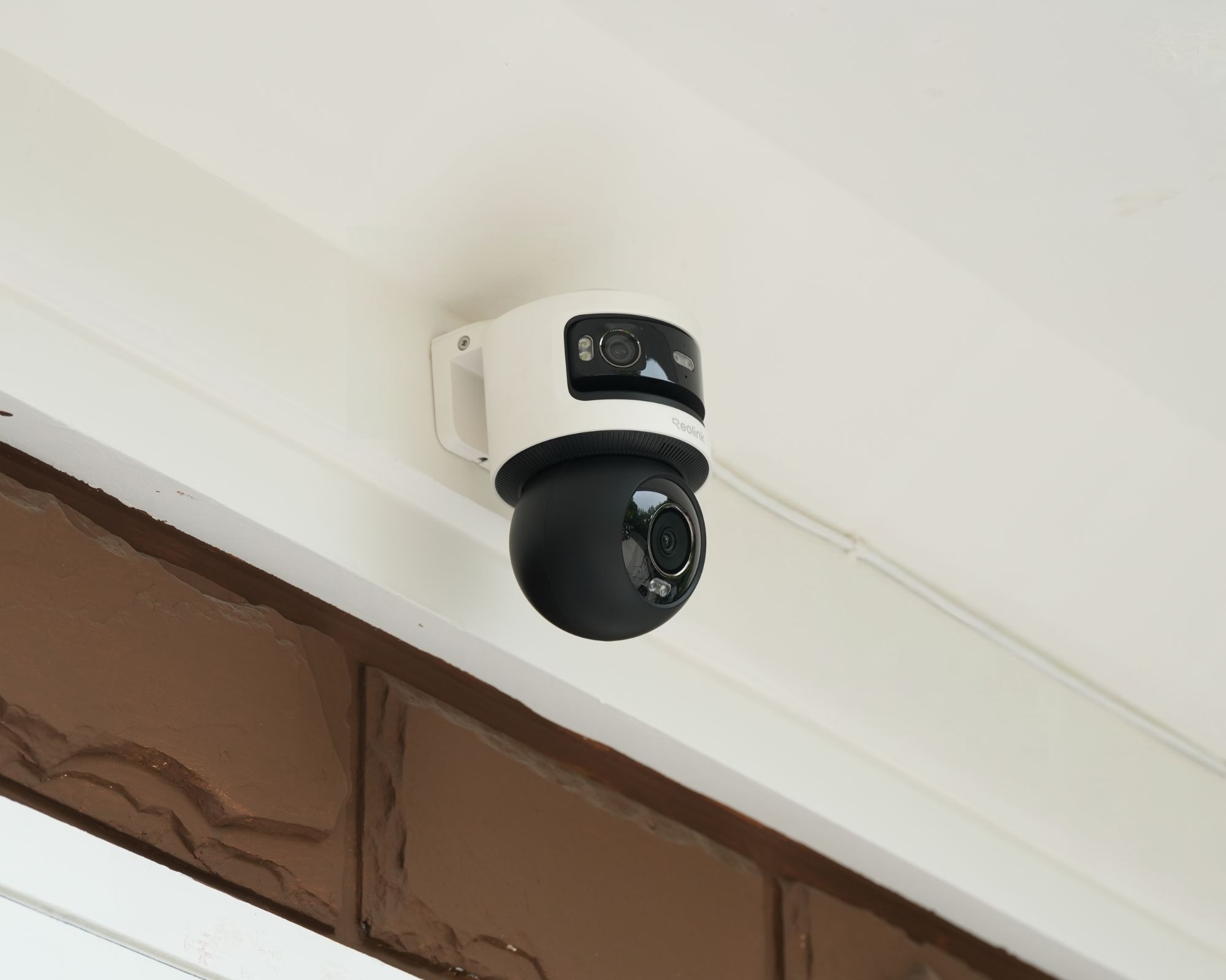 Black and white Reolink OMVI 3i POE installed on a white wall with brown bricks and a white doorframe below.