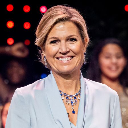 Queen Maxima wearing a blue tie-neck blouse and skirt smiling on stage