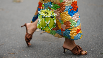 : A guest wears a multicolored print pattern quilted oversized belted long jacket, a green shiny leather with green / white / black pearls straps handbag, brown puffy satin / open toe-cap heels mules , outside Helmstedt, during the Copenhagen Fashion Week Spring/Summer 2024 on August 10, 2023 in Copenhagen, Denmark. 