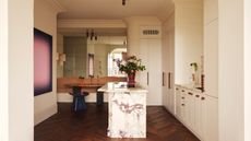 Image of a neutral/cream kitchen with dark hardwood flooring, a dining nook on the back wall, and a pink and white marble island. There is a hot pink painting on the wall.
