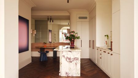 Image of a neutral/cream kitchen with dark hardwood flooring, a dining nook on the back wall, and a pink and white marble island. There is a hot pink painting on the wall.