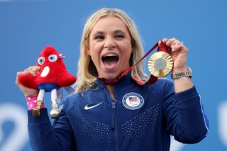 Oksana Masters holds up a gold medal and Paris Phyrge plushy after winning a gold medal at the 2024 Paris Paralympics. She is wearing a navy Team USA sweatsuit and her hair is down and behind her shoulders. She is smiling.